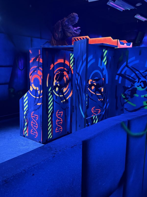 A T-Rex figure over neon-edged bunkers at the arena entrance, lit under black light