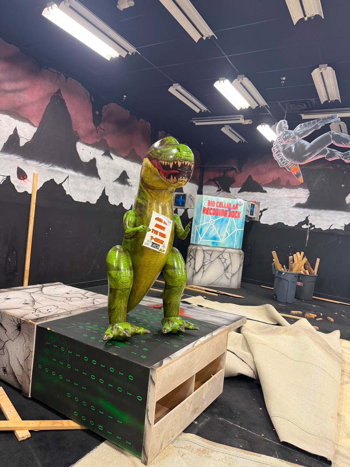 Tye the T-Rex standing on a base in the half-demolished arena, holding an 'Ask us about Tye' sign